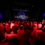 Auf der Bühne spielt ein Orchester. Zuschauer werden rot angestrahlt. Perspektive: Mittig, Hinten.