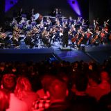 Auf der Bühne spielt ein Orchester. Zuschauer werden rot angestrahlt. Perspektive: Mittig seitlich.