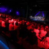 Auf der Bühne spielt ein Orchester. Zuschauer werden rot angestrahlt. Perspektive: Hinten Seite.