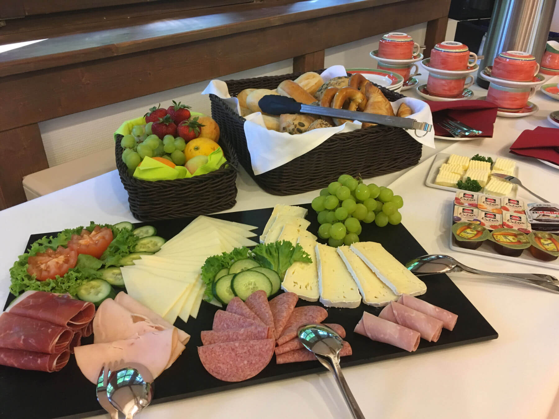 Aufschnitt wie Käse und Wurst, Konfitüre und Brötchen. Dahinter Kaffeetassen.