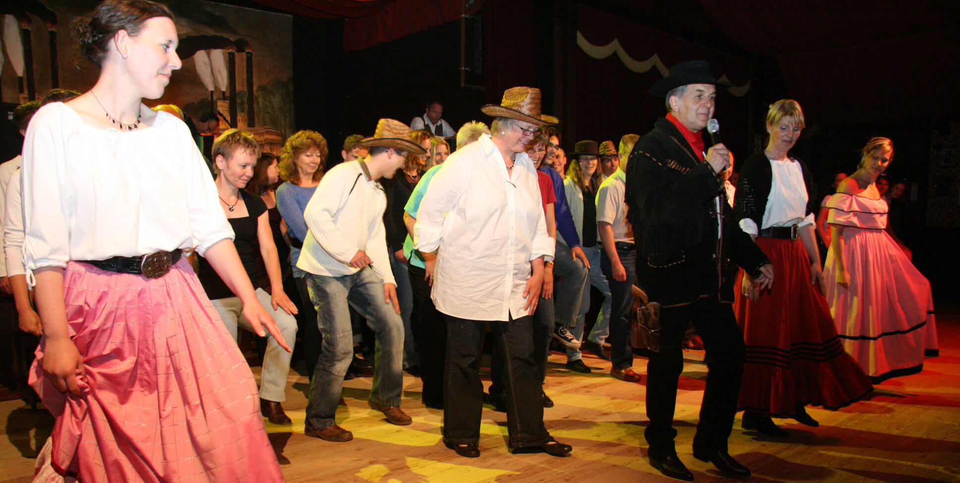 Mann mit Mikrofon tanzt mit Teilnehmern einen Line-Dance. Teilnehmer sind in Reihen dahinter.